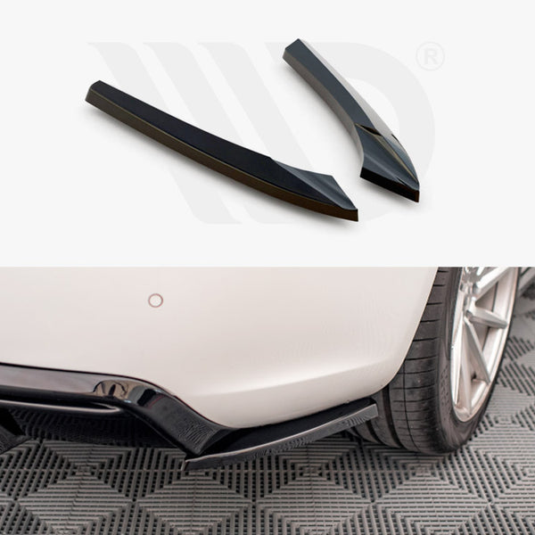 Two black car diffuser fins rest on a white surface above a close-up of a car's rear bumper with a diffuser installed, parked on a patterned grey floor.