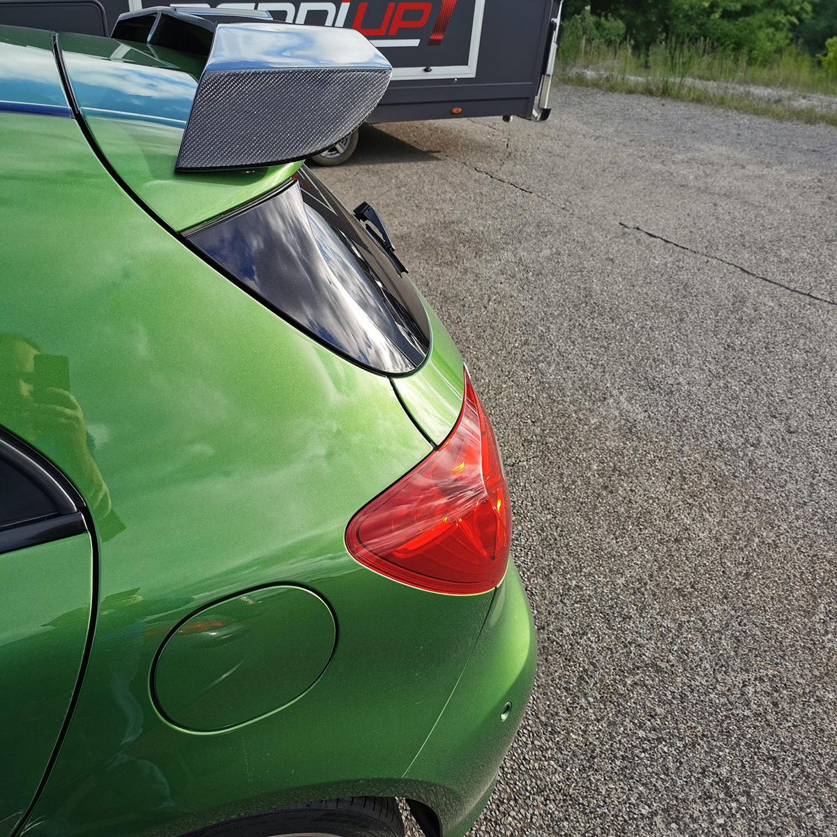 A green car with a carbon fiber spoiler is parked on a gravel surface. In the background, there's a trailer with partial text "UP1".