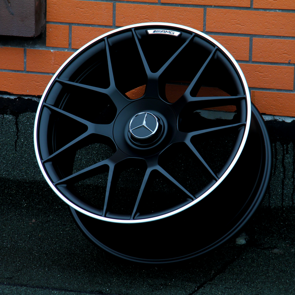 The black AMG-branded car wheel rests against an orange brick wall on a dark pavement. It features a Mercedes emblem in the center.