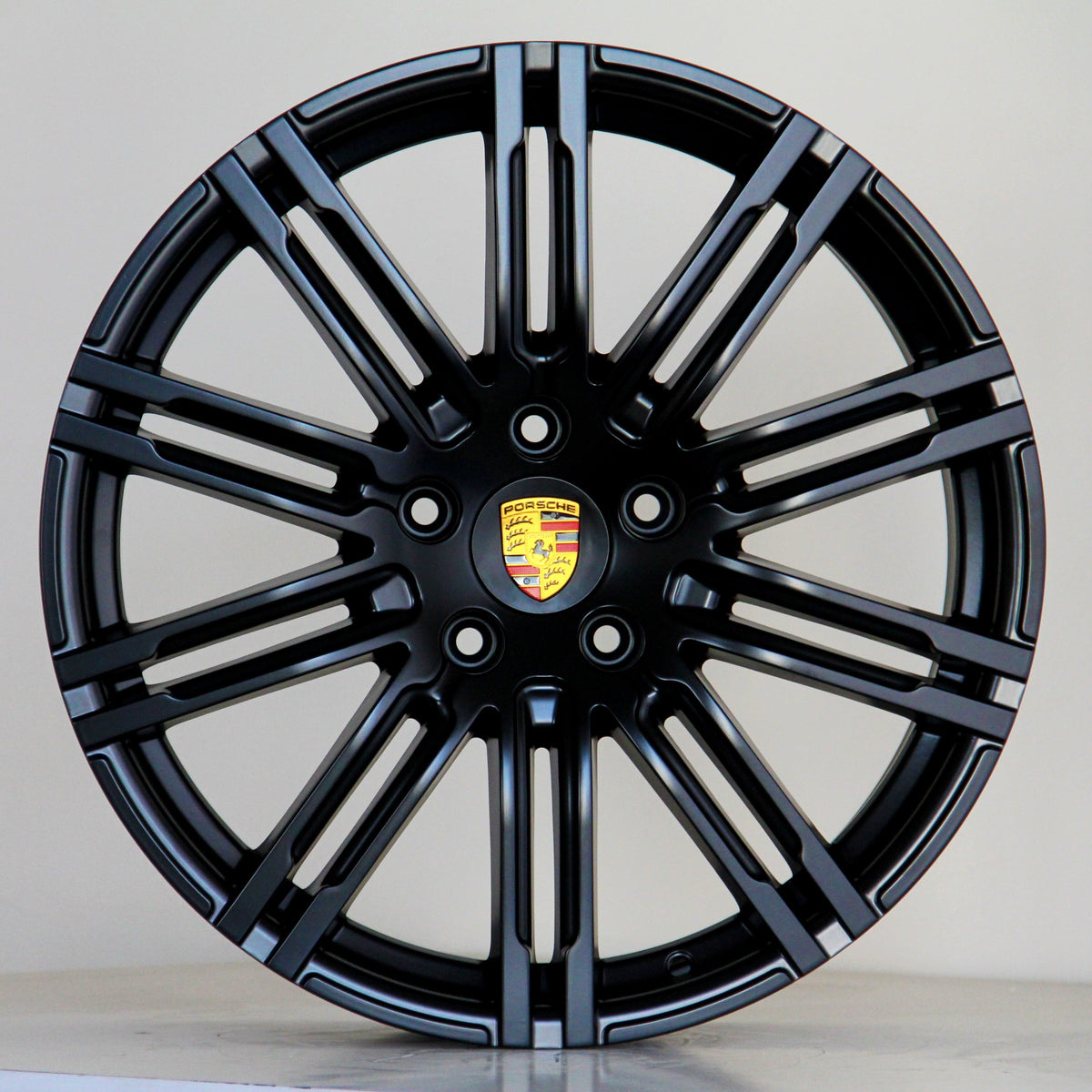 A black alloy wheel with multiple spokes features a central emblem displaying the Porsche logo. The wheel is positioned against a plain light gray background.