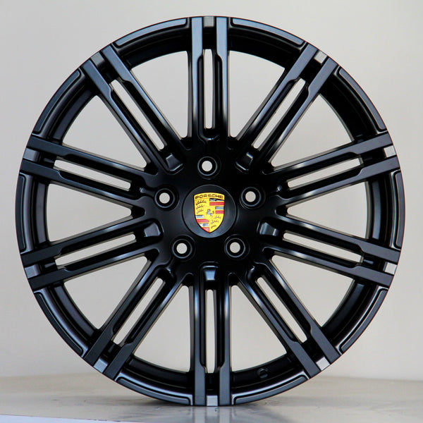 A black alloy wheel with multiple spokes features a central emblem displaying the Porsche logo. The wheel is positioned against a plain light gray background.