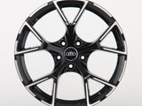 A black alloy wheel with a shiny finish features the Audi logo at its center, set against a plain white background. The wheel has multiple spokes and a sporty design.