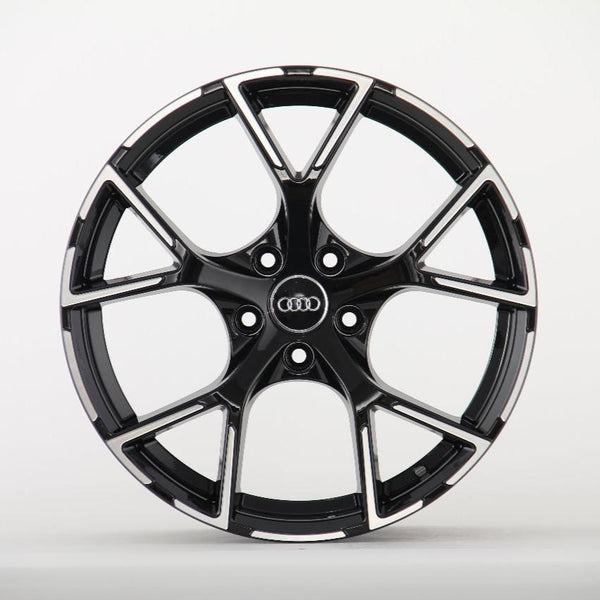 A black alloy wheel with a shiny finish features the Audi logo at its center, set against a plain white background. The wheel has multiple spokes and a sporty design.