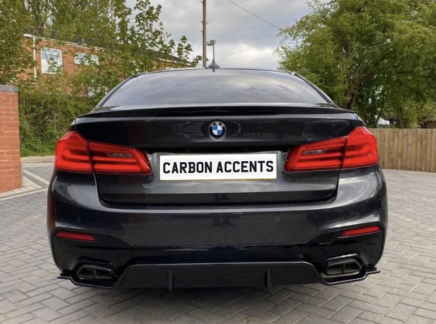 A black BMW sedan is parked on a paved driveway with trees and a brick building in the background. The license plate reads "CARBON ACCENTS."