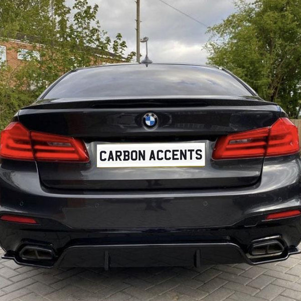 A black BMW sedan is parked on a paved driveway with trees and a brick building in the background. The license plate reads "CARBON ACCENTS."
