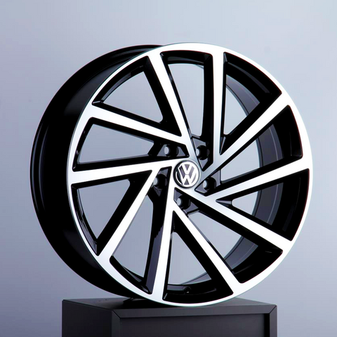 A sleek, modern Volkswagen wheel rim featuring bold geometric spokes is displayed on a black pedestal against a neutral background. It has a glossy black and silver finish with the Volkswagen logo at the center.