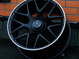 A black Mercedes-Benz AMG wheel leans against an orange brick wall. The wheel features a silver rim and the Mercedes emblem at its center.