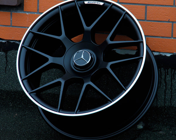 A black Mercedes-Benz AMG wheel leans against an orange brick wall. The wheel features a silver rim and the Mercedes emblem at its center.