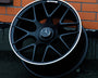 A black Mercedes-Benz AMG wheel leans against an orange brick wall. The wheel features a silver rim and the Mercedes emblem at its center.