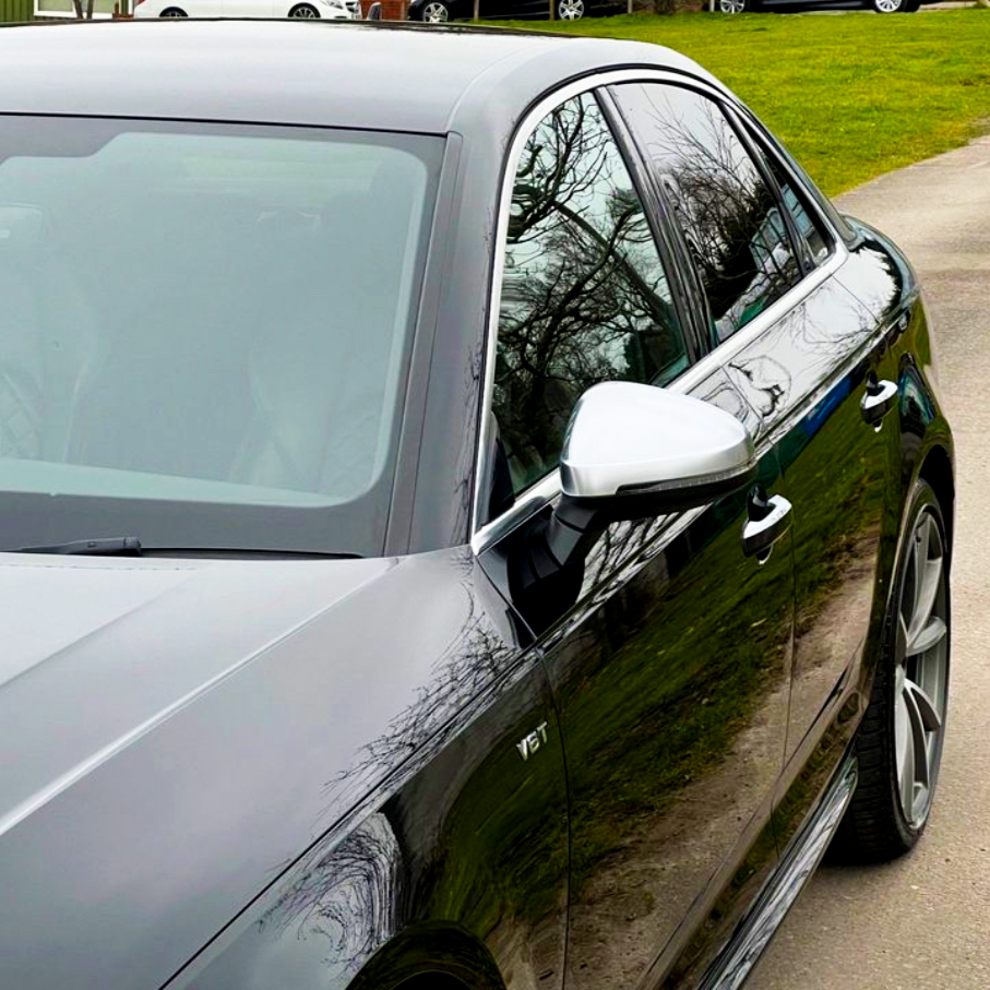 A shiny black car parked on a driveway, reflecting trees in its sleek surface. The side mirror is visible, with green grass and other cars in the background. The text "V6T" is visible on the side.