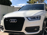 A white Audi SUV is parked on a paved area under a sunny sky. The front grille features the Audi logo, surrounded by a residential fence and trees.