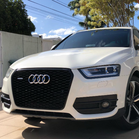 A white Audi SUV is parked on a paved area under a sunny sky. The front grille features the Audi logo, surrounded by a residential fence and trees.