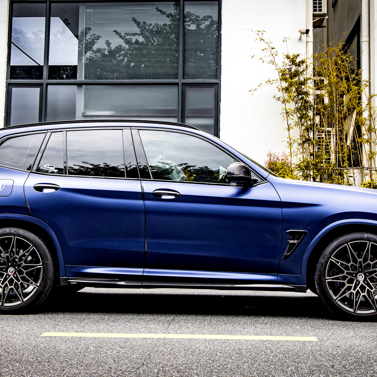 A blue SUV is parked on a street beside a modern building with large windows and greenery, featuring intricate wheel design and sleek body lines.