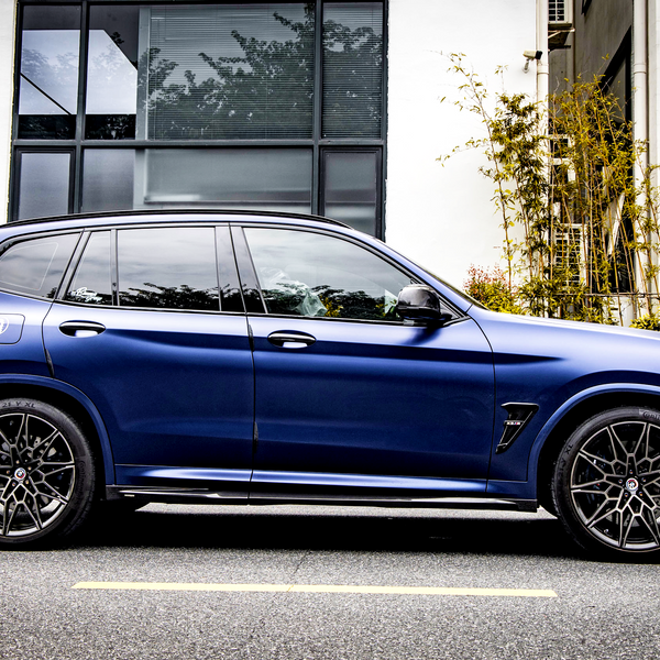 A blue SUV is parked on a street beside a modern building with large windows and greenery, featuring intricate wheel design and sleek body lines.