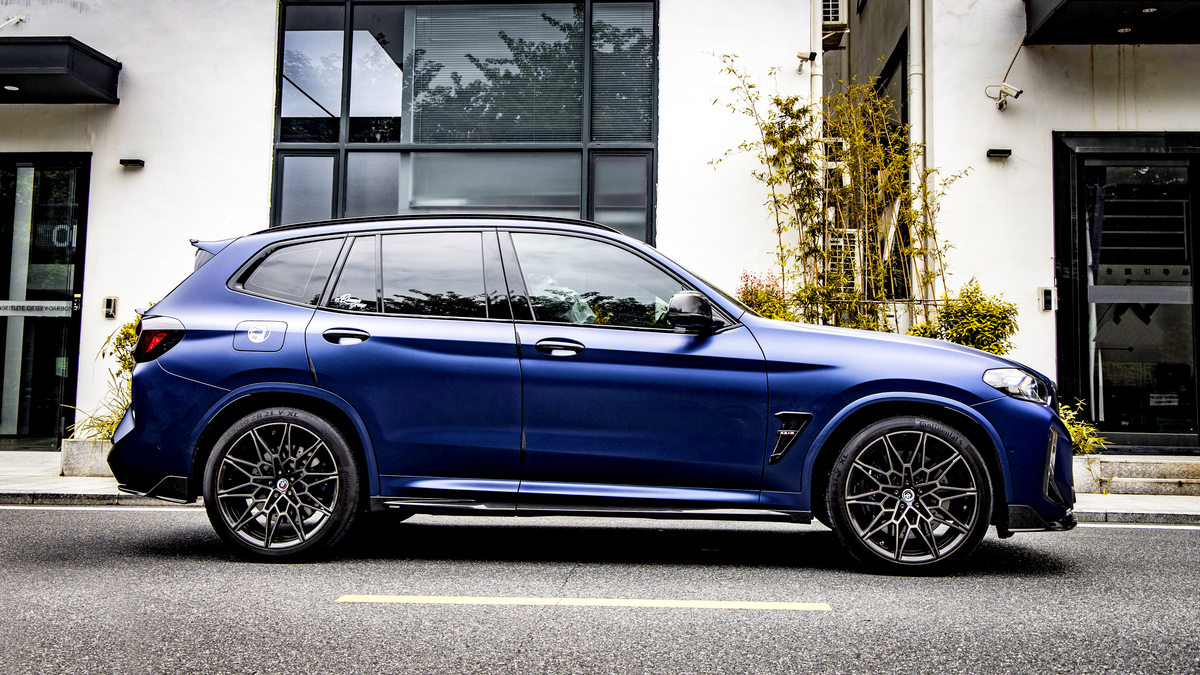 A blue SUV is parked on a street beside a modern building with large windows and greenery, featuring intricate wheel design and sleek body lines.