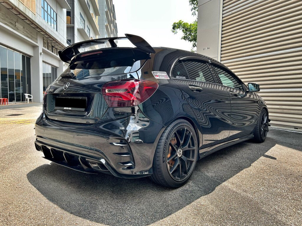A black Mercedes hatchback, parked beside industrial buildings, features dark-tinted windows, a rear spoiler, and alloys. Reflective surfaces display building textures. Visible logos: "AMG" and "Ecobenzo."