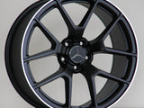 A black alloy wheel with a Mercedes-Benz logo is resting on a wooden surface against a plain background. Text on the rim reads 