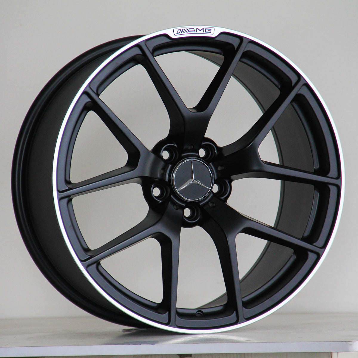 A black alloy wheel with a Mercedes-Benz logo is resting on a wooden surface against a plain background. Text on the rim reads "AMG" with a stylized logo.