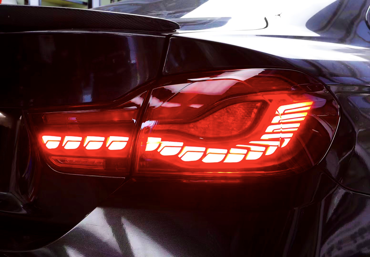 A car's taillight is illuminated in bright red, showing a sleek, modern design with horizontal light strips, set against a glossy black car exterior.