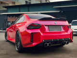 A red BMW M2 car is parked on a concrete surface, facing away, showing its sleek rear design and quad exhausts. It's set against an industrial building backdrop.