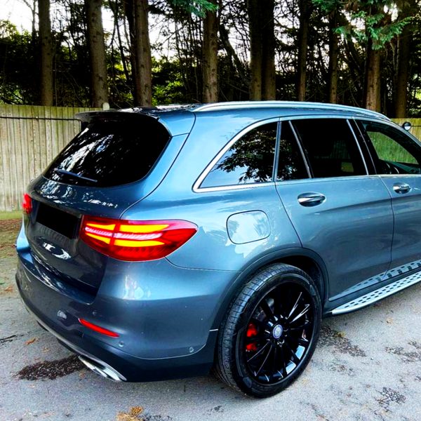 A grey SUV is parked on a paved area, surrounded by tall trees and a wooden fence, with its taillights illuminated.