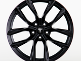 A black Tesla wheel rim with a sleek, multi-spoke design is shown in a clean, white studio setting. The central hub cap features the Tesla logo prominently.
