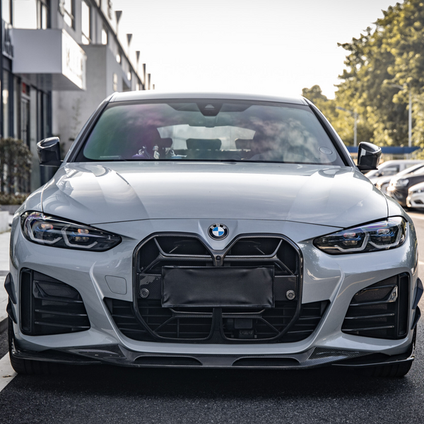 A silver car with a prominent grille is parked on a street beside a line of buildings, with trees visible in the background under a clear sky.