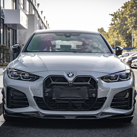 A silver car with a prominent grille is parked on a street beside a line of buildings, with trees visible in the background under a clear sky.