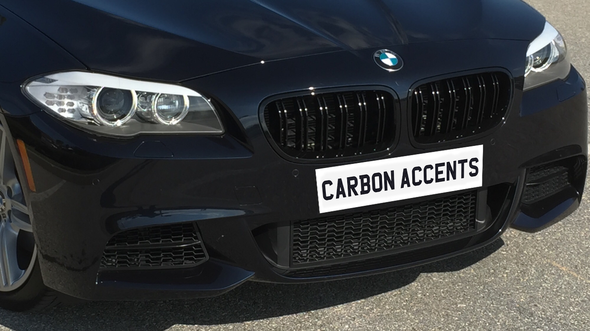A black car features carbon accents on its front grille, parked on a paved surface. Text on the license plate area reads "CARBON ACCENTS."