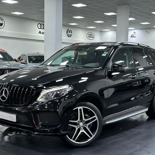 A black Mercedes SUV is parked inside a car showroom, surrounded by other vehicles. Logos of BMW, Audi, Volkswagen, and Land Rover are displayed on the walls.