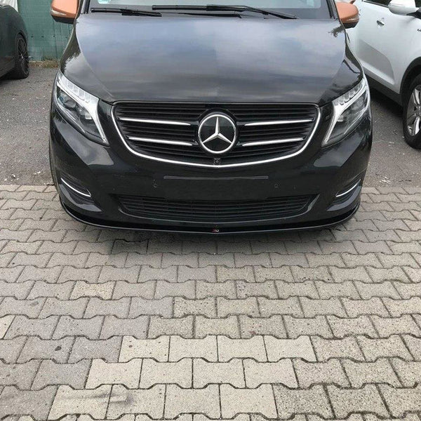 A black Mercedes-Benz van is parked on a paved area, surrounded by other cars, including a white SUV on the right and a dark vehicle on the left.