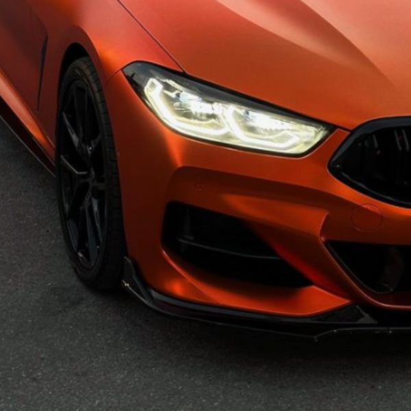 Car's front end with bright headlights illuminated; matte orange finish and black grille. Parked on a dark asphalt surface, displaying a sporty and sleek design.