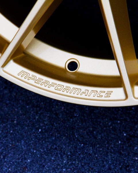 A golden car wheel rim engraved with "MPERFORMANCE" rests on a dark, textured ground.