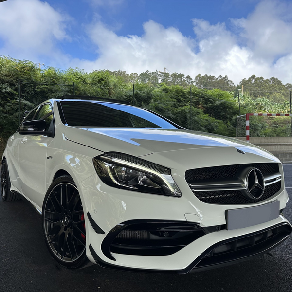 A white Mercedes-Benz car is parked on a dark paved area under a clear, blue sky, adjacent to greenery and a red-and-white soccer goalpost.