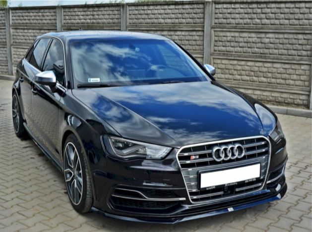 A sleek black Audi car, parked on a cobblestone driveway, reflects the cloudy blue sky. It is situated in front of a tall, textured stone wall.
