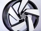 A car wheel rim displays a black and silver geometric design featuring a central Volkswagen logo. It is positioned upright against a neutral, softly lit background.