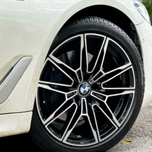 A car's modern alloy wheel with a BMW logo is stationary on a paved road. The surrounding area is part of a white vehicle's side body.