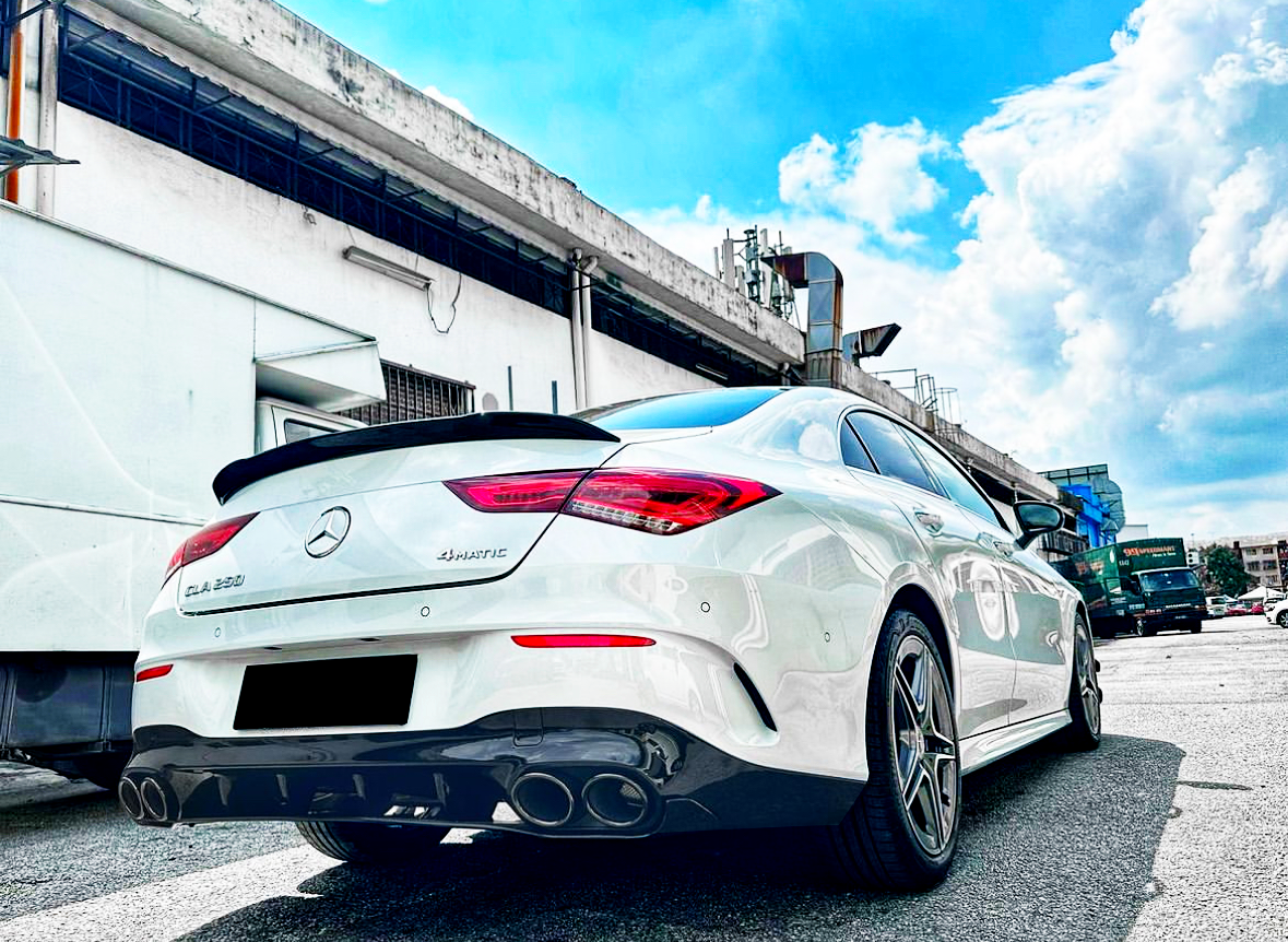 Car parked on a street next to a building under a clear blue sky. The white vehicle is a Mercedes CLA 250 4MATIC, with dual exhaust pipes and silver alloy wheels.