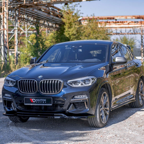 A black BMW SUV is parked on a gravel path, with metal scaffolding and green foliage in the background. The license plate reads "MAXTON design."
