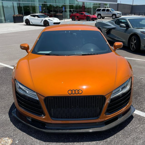 Orange Audi sports car parked in a lot, surrounded by several other high-end cars, including a white and a gray vehicle, outside a glass-fronted building.