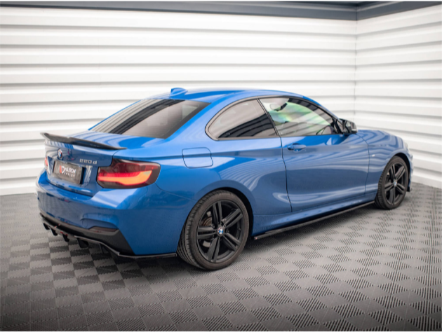 A blue sports car with black wheels is parked in a well-lit, enclosed showroom. The vehicle has a visible badge, "220d," on the rear, indicating its model.