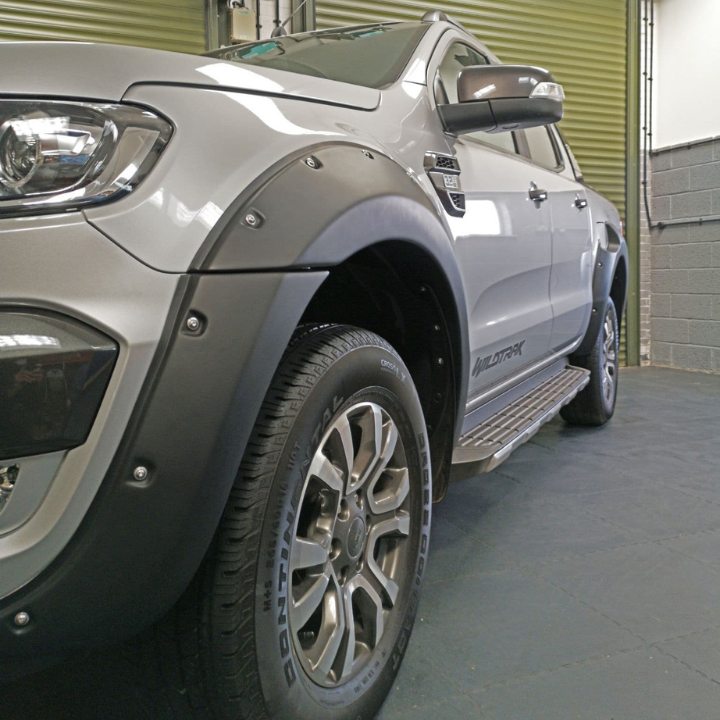 A silver pickup truck parked inside a garage with green roll-up doors visible. "Wildtrak" is written on the side. The image highlights the front wheel and fender.
