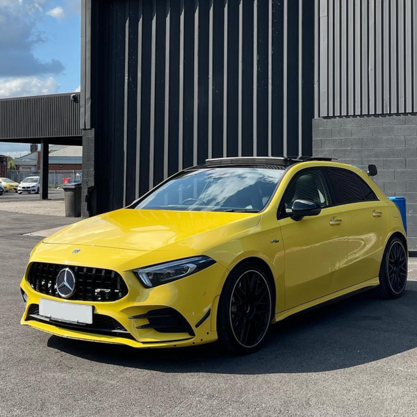 A yellow Mercedes-Benz car is parked on an asphalt surface, set against a large gray industrial building and a partly cloudy sky.