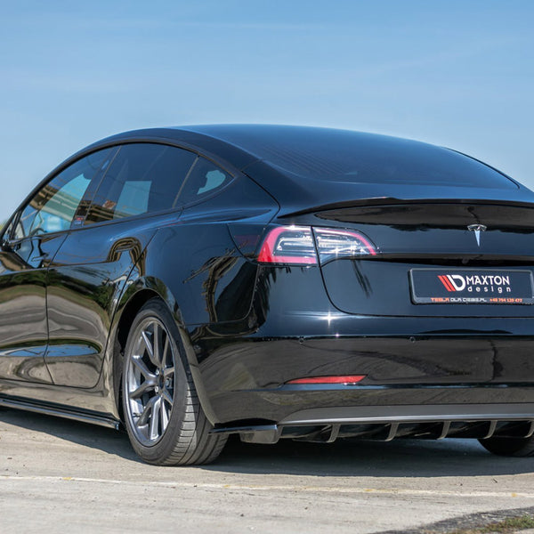 A black car parked on a pavement, showcasing its sleek design with tinted windows and alloy wheels. The background includes a clear blue sky and distant buildings. License plate reads: Maxton Design.