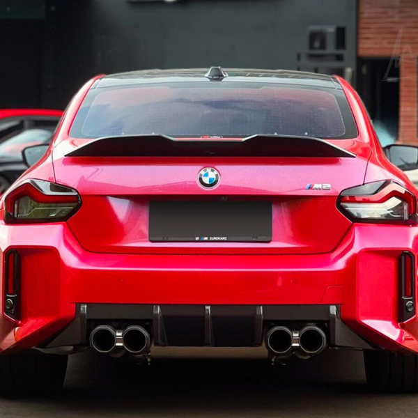A bright red BMW M2 car is parked, displaying quad exhaust pipes. It is surrounded by other parked vehicles in an outdoor setting. Text on plate reads, "EUROKARS."