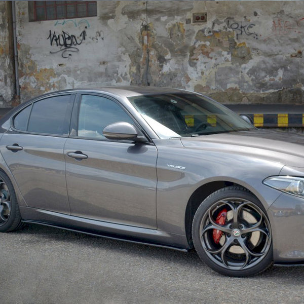A silver car parked beside a weathered wall covered in graffiti and peeling paint. The ground is paved, and the word "VELOCE" is visible on the car's side.