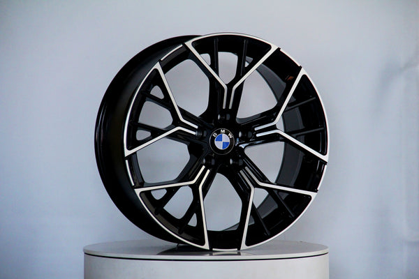 A black and silver BMW wheel rim stands vertically on a circular white platform against a plain, light background. The BMW logo is centered on the wheel.
