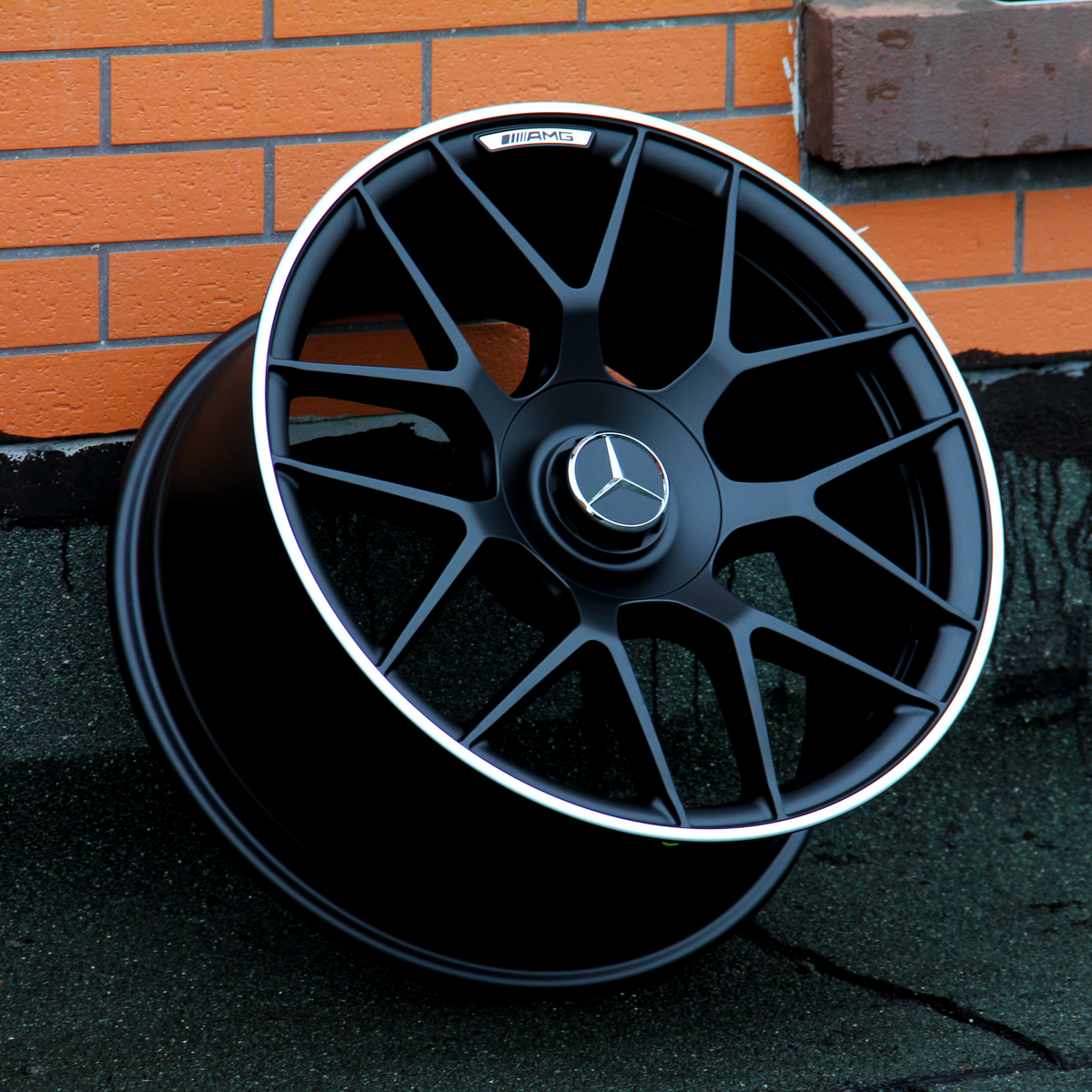 A black Mercedes wheel with a star emblem and AMG label leans against a brick wall on a rough concrete surface.