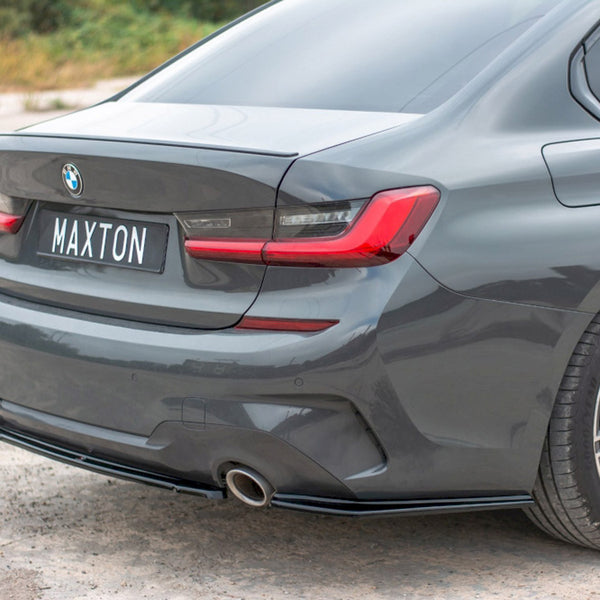 A grey BMW car is parked, showcasing its rear with a "MAXTON" license plate. The setting is an open, paved outdoor area with greenery in the background.