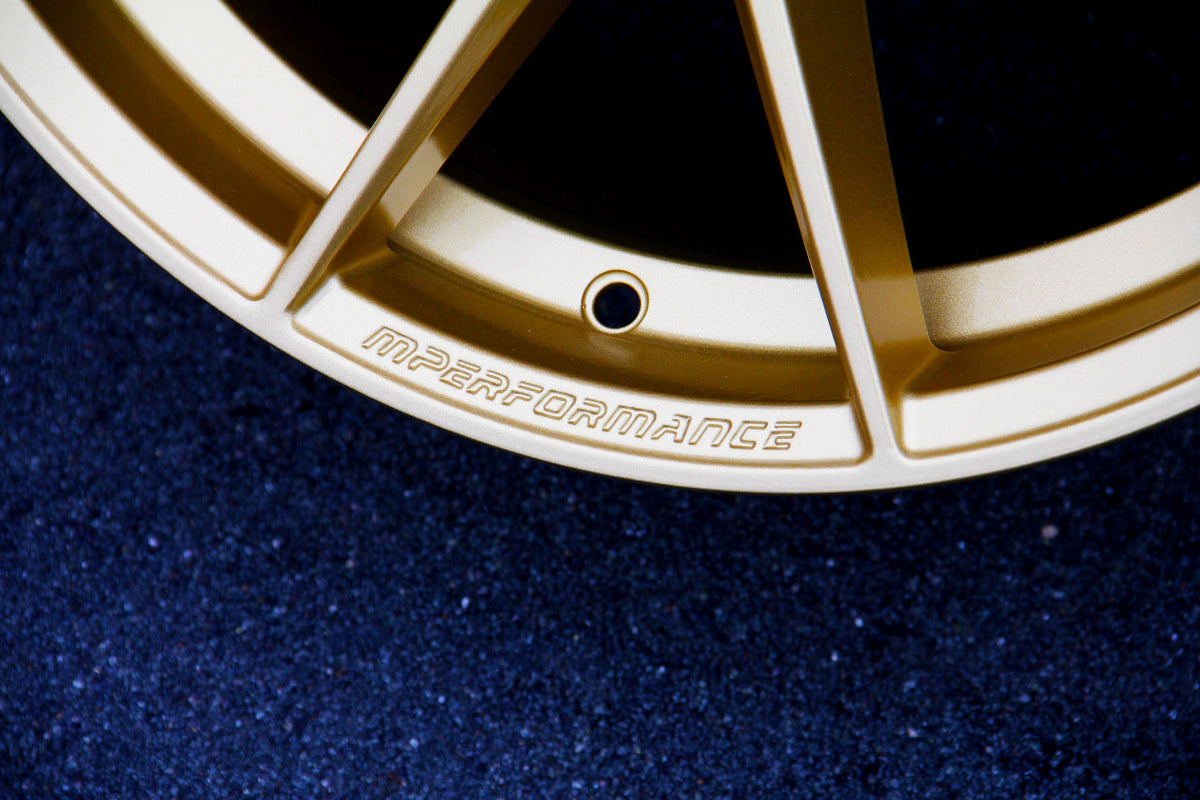 A golden wheel rim with the text "MPERFORMANCE" engraved along the edge, resting on a textured dark surface.